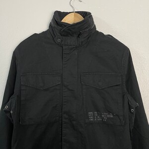 May include: Black military-style jacket with a high collar and multiple pockets. The jacket has a button closure and a zipper detail on the collar. The garment is hanging on a metal hanger against a plain white background. Text is printed on the right side of the jacket.