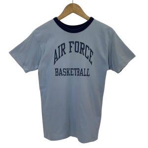 May include: A light blue short-sleeve t-shirt with a dark blue crew neckline. The front features the text AIR FORCE in an arched font above the word BASKETBALL in a straight font, both printed in dark blue ink on the chest of the garment.