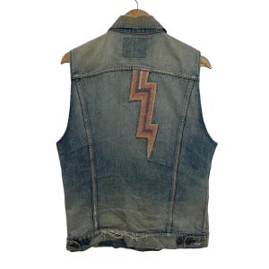 May include: A light blue denim vest with a large embroidered lightning bolt on the back. The lightning bolt is made of pink, orange, and blue thread.