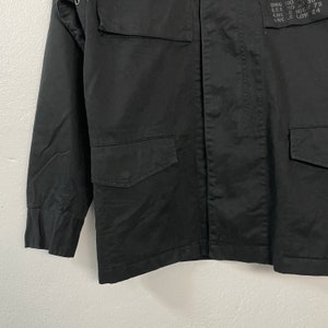May include: Black military-style jacket with four front pockets and a button-up closure. The jacket has long sleeves and a collar. Small text is visible on the upper right side. The jacket is made of a durable fabric.