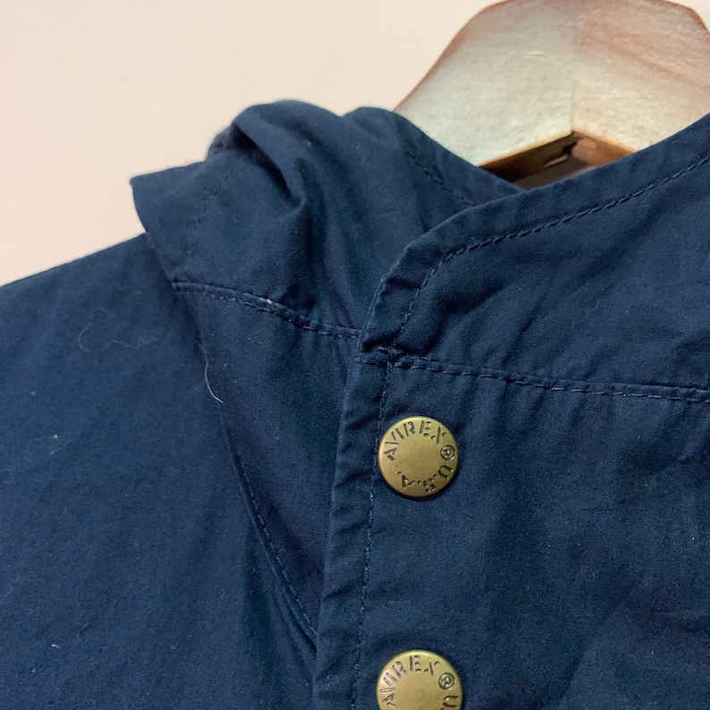 May include: A close-up shot of a navy blue jacket collar featuring two brass-coloured buttons. The buttons are embossed with the text "AVIREX U.S.A." The jacket appears to be made of a smooth fabric. The collar is folded over, and the jacket is hanging on a wooden hanger.
