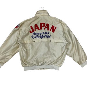 May include: Cream-colored vintage jacket with the word "JAPAN" in red block letters across the back. Below the word "JAPAN" is the text "History of JKA KANAGAWA" in blue. The jacket has a white ribbed collar, cuffs, and waistband.