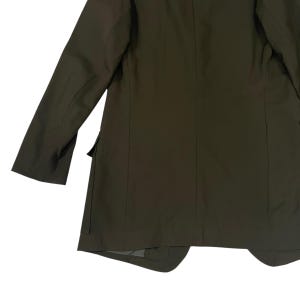 Puede incluir: Una americana de color verde oliva con mangas largas, cuello estructurado y un bolsillo lateral con solapa. La prenda est&aacute; confeccionada con costuras verticales en la espalda y los costados, mostrando un acabado de tela lisa y uniforme.