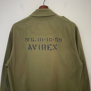 Op de afbeelding: Olijfgroene jas met de tekst "MFG. 10-16-59 AVIREX" in zwart op de rug gedrukt. De jas heeft een kraag en lange mouwen. Hij hangt aan een zwarte hanger tegen een witte achtergrond.