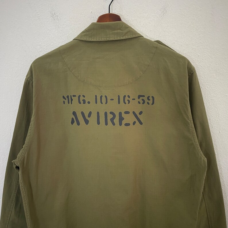 Op de afbeelding: Olijfgroene jas met de tekst "MFG. 10-16-59 AVIREX" in zwart op de rug gedrukt. De jas heeft een kraag en lange mouwen. Hij hangt aan een zwarte hanger tegen een witte achtergrond.