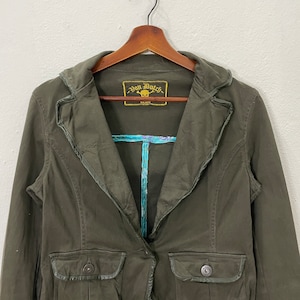 May include: A dark green, long-sleeved jacket with a single button closure. The jacket has a collar with a decorative trim and a pocket on the left side. The label on the jacket reads "Von Dutch Est. 1972".