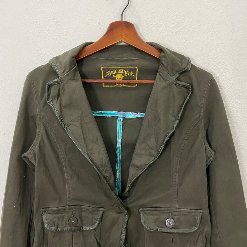 May include: A dark green, long-sleeved jacket with a single button closure. The jacket has a collar with a decorative trim and a pocket on the left side. The label on the jacket reads "Von Dutch Est. 1972".
