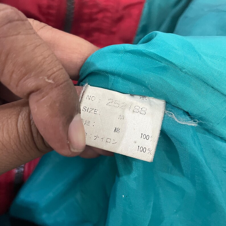 K&ouml;nnte beinhalten: Nahaufnahme eines wei&szlig;en Etiketts mit japanischem Text und der Nummer "282188". Das Etikett ist an einem t&uuml;rkisfarbenen Stoff befestigt, der Teil eines Kleidungsst&uuml;cks mit roten und t&uuml;rkisfarbenen Abschnitten ist.