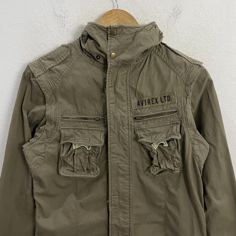 May include: Olive green jacket with multiple pockets and a high collar. The jacket features the text "AVIREX LTD" on a patch above the chest pocket. The jacket is hanging on a wooden hanger.