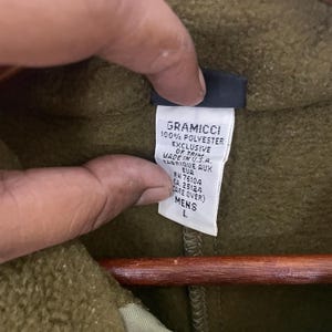 May include: Close-up of a tag from a Gramicci jacket. The tag is white with black text, indicating it's made of 100% polyester and made in the U.S.A. The size is listed as L, and the style is for men. The jacket is olive green.