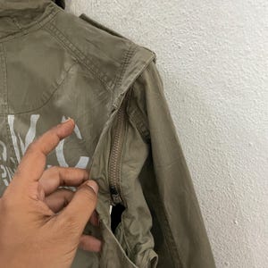 May include: Olive green jacket with a zipper closure and white lettering. The jacket has a hood and a pocket. The fabric appears to be lightweight and slightly wrinkled. The jacket is a casual style.