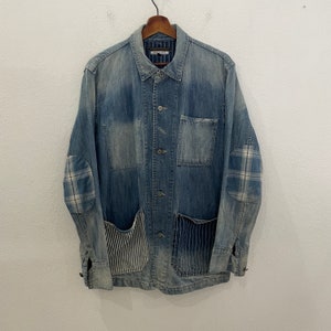 May include: A blue denim jacket with patchwork details, including plaid elbow patches and striped fabric pockets. The jacket has a button-up front and long sleeves.