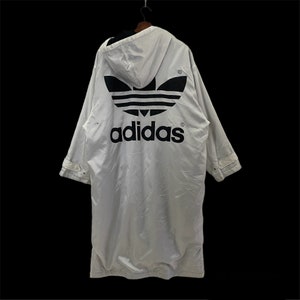 May include: White Adidas long coat with a hood and black logo on the back. The Adidas logo is a large trefoil with the brand name below it. The coat has long sleeves and a button closure.