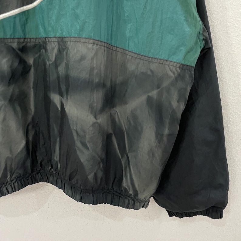 May include: A vintage windbreaker jacket in black, teal, and gray. The jacket features a color-blocked design with a drawstring at the hood and a cinched waist. The material has a slight sheen, suggesting it's made of nylon or a similar fabric.