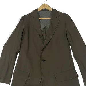 Puede incluir: Un blazer de color verde oliva s&oacute;lido con solapa de muesca, cierre frontal de un solo bot&oacute;n y dos bolsillos con solapa en la cintura. La prenda se muestra en una percha de madera e incluye un forro interior gris visible en el cuello.