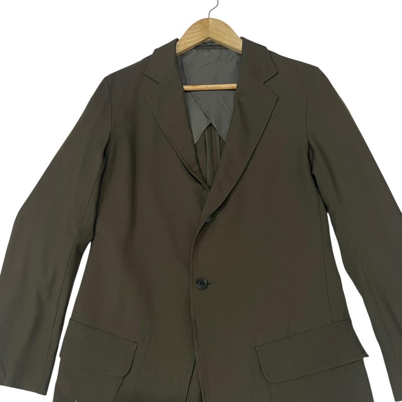 Puede incluir: Un blazer de color verde oliva s&oacute;lido con solapa de muesca, cierre frontal de un solo bot&oacute;n y dos bolsillos con solapa en la cintura. La prenda se muestra en una percha de madera e incluye un forro interior gris visible en el cuello.