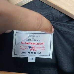 May include: Close-up of a black jacket's interior, showcasing a white label with the text "Certificate of Authenticity" and "All American Classic." The label includes the American flag and the brand name "AVIREX U.S.A." The size "M" is also visible.