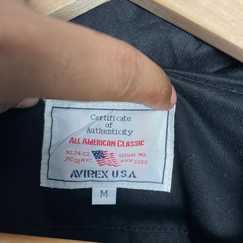 May include: Close-up of a black jacket's interior, showcasing a white label with the text "Certificate of Authenticity" and "All American Classic." The label includes the American flag and the brand name "AVIREX U.S.A." The size "M" is also visible.