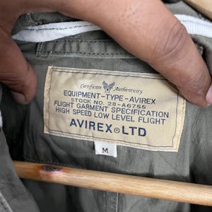 May include: Close-up of an olive green jacket's interior, featuring a label with text: "EQUIPMENT-TYPE-AVIREX, STOCK NO. 28-A6755, FLIGHT GARMENT SPECIFICATION, HIGH SPEED LOW LEVEL FLIGHT, AVIREX LTD." The label also has a size "M".