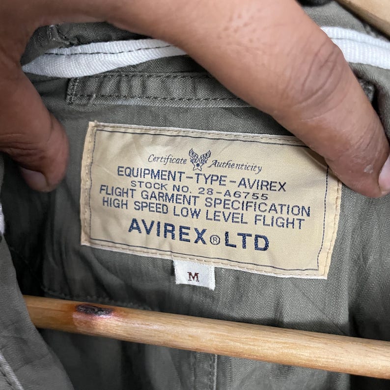 May include: Close-up of an olive green jacket's interior, featuring a label with text: "EQUIPMENT-TYPE-AVIREX, STOCK NO. 28-A6755, FLIGHT GARMENT SPECIFICATION, HIGH SPEED LOW LEVEL FLIGHT, AVIREX LTD." The label also has a size "M".