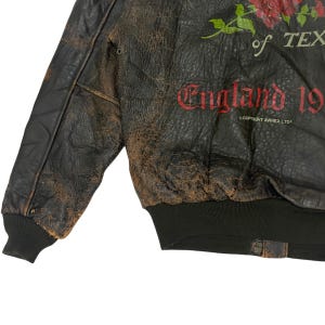 May include: A distressed dark brown leather bomber jacket featuring a red rose graphic, the text of TEXAS, and England 19. The jacket includes a black ribbed hem and cuffs, with the small printed text COPYRIGHT AVIREX LTD visible on the front.