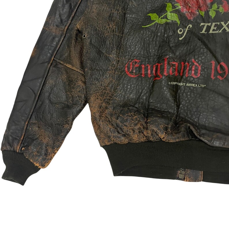 May include: A distressed dark brown leather bomber jacket featuring a red rose graphic, the text of TEXAS, and England 19. The jacket includes a black ribbed hem and cuffs, with the small printed text COPYRIGHT AVIREX LTD visible on the front.