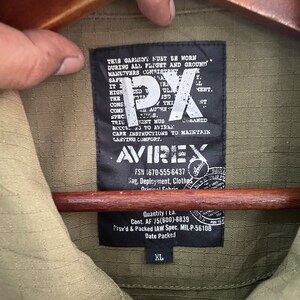 Op de afbeelding: Close-up van een olijfgroene jas in militaire stijl met een zwart label. Het label toont het merk "AVIREX" en de tekst "PX". De jas is maat XL.