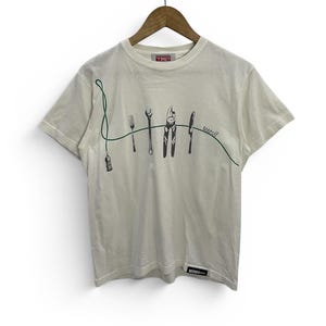 May include: A cream-coloured short-sleeve t-shirt featuring a graphic print of a green line connecting a corkscrew, fork, wrench, pliers, and knife. The text "Merci!" is printed in green near the knife. A small rectangular brand tag is at the hem.