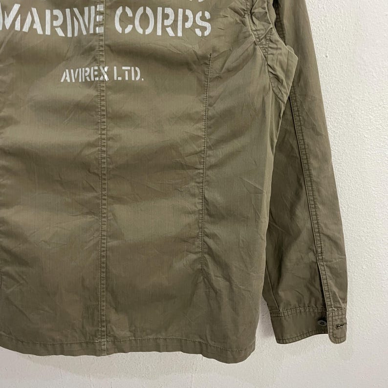 May include: Olive green jacket with the text "MARINE CORPS" and "AVIREX LTD." printed in white. The jacket has a buttoned cuff and a classic military style.