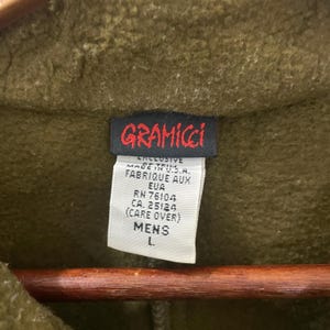 May include: Close-up of a green Gramicci jacket tag. The tag is black with red lettering and white text. The tag reads "GRAMICCI" and includes details about the garment's origin and care instructions. The size is listed as "L."