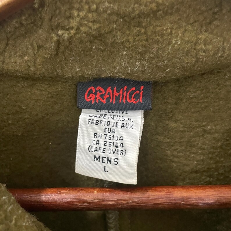 May include: Close-up of a green Gramicci jacket tag. The tag is black with red lettering and white text. The tag reads "GRAMICCI" and includes details about the garment's origin and care instructions. The size is listed as "L."