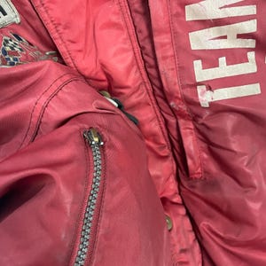 K&ouml;nnte beinhalten: Nahaufnahme einer roten Jacke mit silbernem Rei&szlig;verschluss und dem wei&szlig;en Aufdruck "TEAM". Die Jacke hat einen kleinen Aufn&auml;her mit einem karierten Flaggenmuster. Die Jacke scheint aus Nylon oder einem &auml;hnlichen Material zu sein.