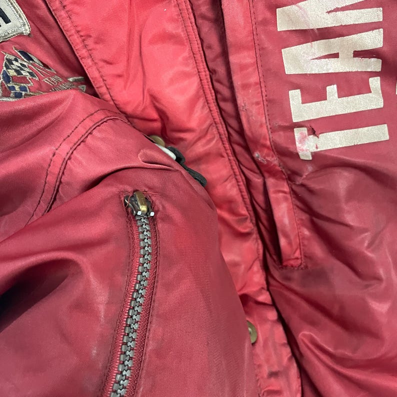 K&ouml;nnte beinhalten: Nahaufnahme einer roten Jacke mit silbernem Rei&szlig;verschluss und dem wei&szlig;en Aufdruck "TEAM". Die Jacke hat einen kleinen Aufn&auml;her mit einem karierten Flaggenmuster. Die Jacke scheint aus Nylon oder einem &auml;hnlichen Material zu sein.