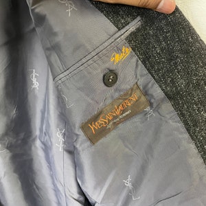 Op de afbeelding: De binnenkant van een grijze blazer met een zwarte knoop en een bruin label met de tekst "Yves Saint Laurent, diffusion homme" in wit.