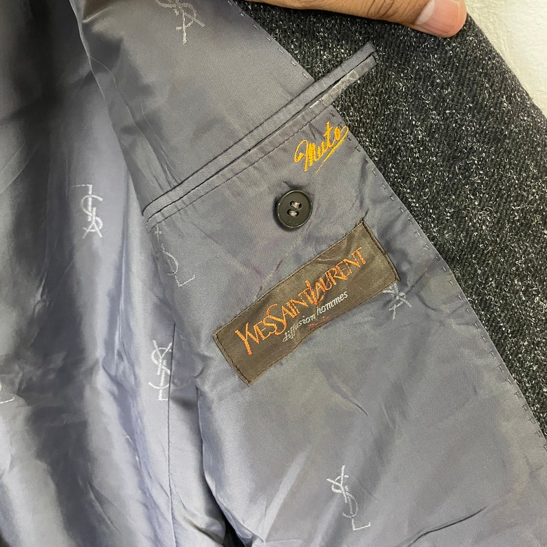 Op de afbeelding: De binnenkant van een grijze blazer met een zwarte knoop en een bruin label met de tekst "Yves Saint Laurent, diffusion homme" in wit.