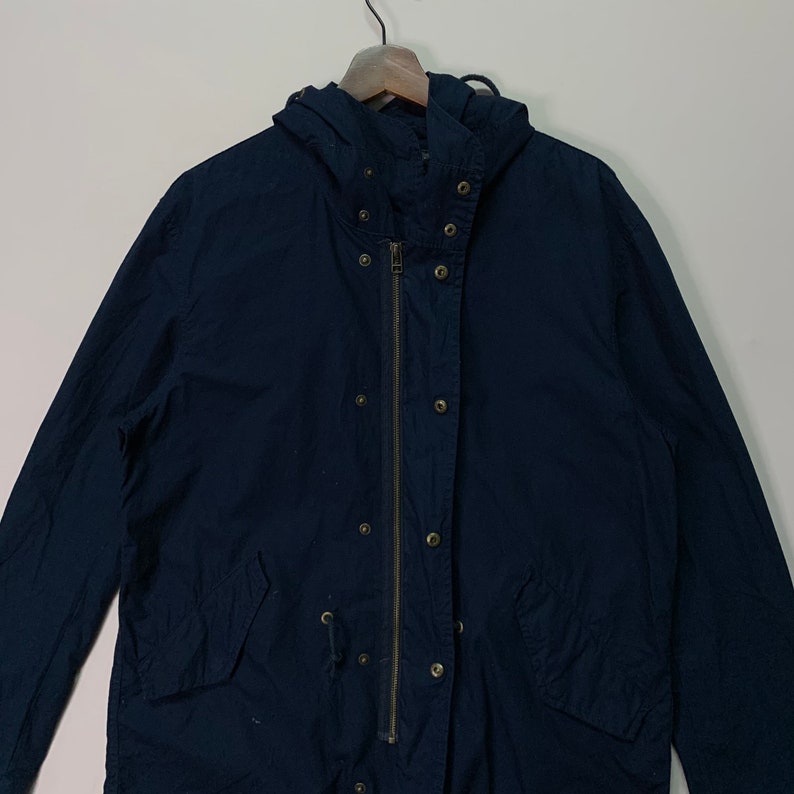 May include: A navy blue hooded jacket with a zip and snap fastenings. The jacket features a drawstring waist and is suspended from a wooden hanger. The jacket is made of a lightweight fabric.