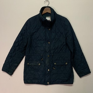 Op de afbeelding: Een donkerblauw gewatteerd jack met een hoge kraag, een taille met trekkoord en twee klepzakken. Het jack heeft een knoopsluiting en goudkleurige knopen.