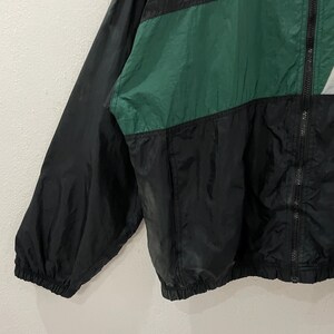 May include: A black, green, and gray nylon windbreaker jacket. The jacket has a full zipper and a color-block design. The sleeves are black, and the body features green and gray panels. The jacket is lightweight and appears to be vintage.