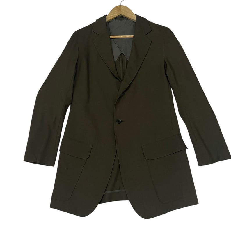 Puede incluir: Un blazer de color marr&oacute;n oscuro con solapa de muesca, cierre frontal de un solo bot&oacute;n y dos bolsillos grandes con solapa en la cintura. La prenda se muestra en una percha de madera, con mangas largas y una silueta estructurada.