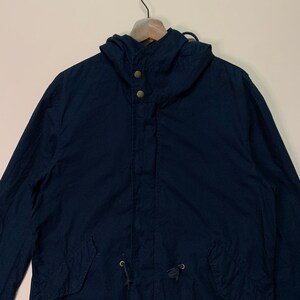 May include: A navy blue hooded jacket with a drawstring waist and two gold-coloured buttons at the collar. The jacket has a hood and two front pockets. The jacket is hanging on a wooden hanger.