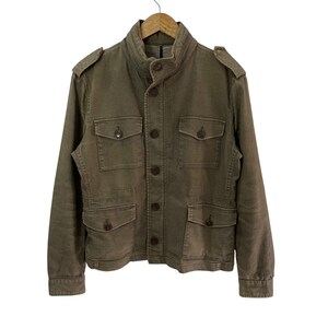 May include: A brown utility jacket featuring a high collar, a front button closure, and four flap pockets on the chest and waist. The jacket includes shoulder epaulets, long sleeves with buttoned cuffs, and a structured, durable fabric construction.
