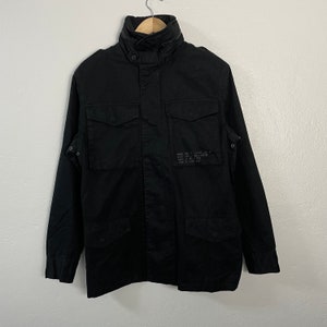 May include: Black military-style jacket with a high collar, two chest pockets, and two lower pockets. The jacket has a button-up front and a small patch of text on the right side. The jacket is hanging on a wooden hanger.