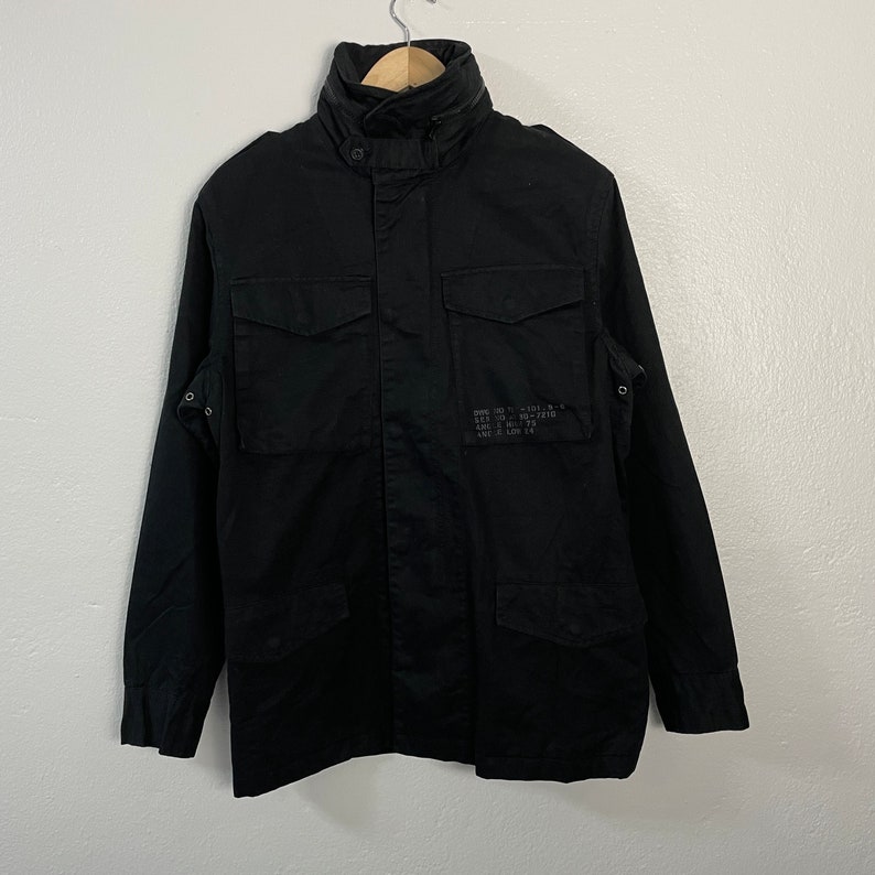 May include: Black military-style jacket with a high collar, two chest pockets, and two lower pockets. The jacket has a button-up front and a small patch of text on the right side. The jacket is hanging on a wooden hanger.