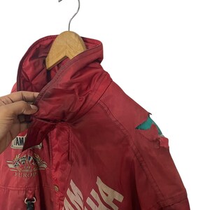 K&ouml;nnte beinhalten: Rote Yamaha-Rennjacke mit wei&szlig;em Schriftzug und Aufn&auml;hern. Die Jacke hat eine Kapuze und einen Rei&szlig;verschluss. Die Jacke h&auml;ngt an einem Holzb&uuml;gel. Abnutzungserscheinungen sind sichtbar.