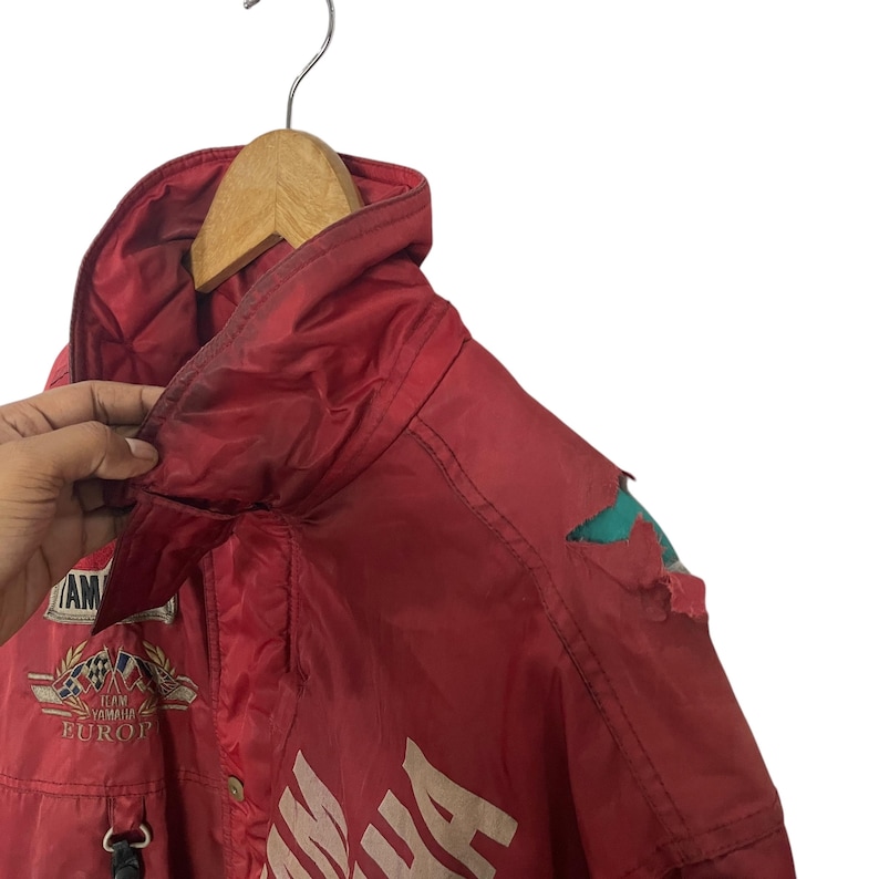 K&ouml;nnte beinhalten: Rote Yamaha-Rennjacke mit wei&szlig;em Schriftzug und Aufn&auml;hern. Die Jacke hat eine Kapuze und einen Rei&szlig;verschluss. Die Jacke h&auml;ngt an einem Holzb&uuml;gel. Abnutzungserscheinungen sind sichtbar.