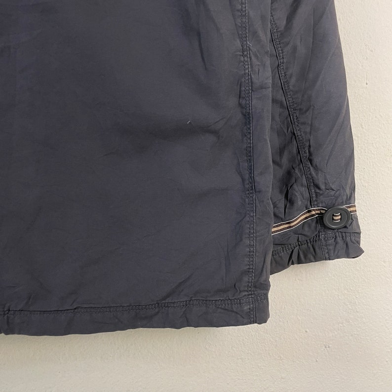 Pode incluir: Close-up de um casaco azul marinho escuro. O casaco tem um fecho de bot&atilde;o e um detalhe de riscas decorativas no punho. O tecido parece leve e ligeiramente enrugado.