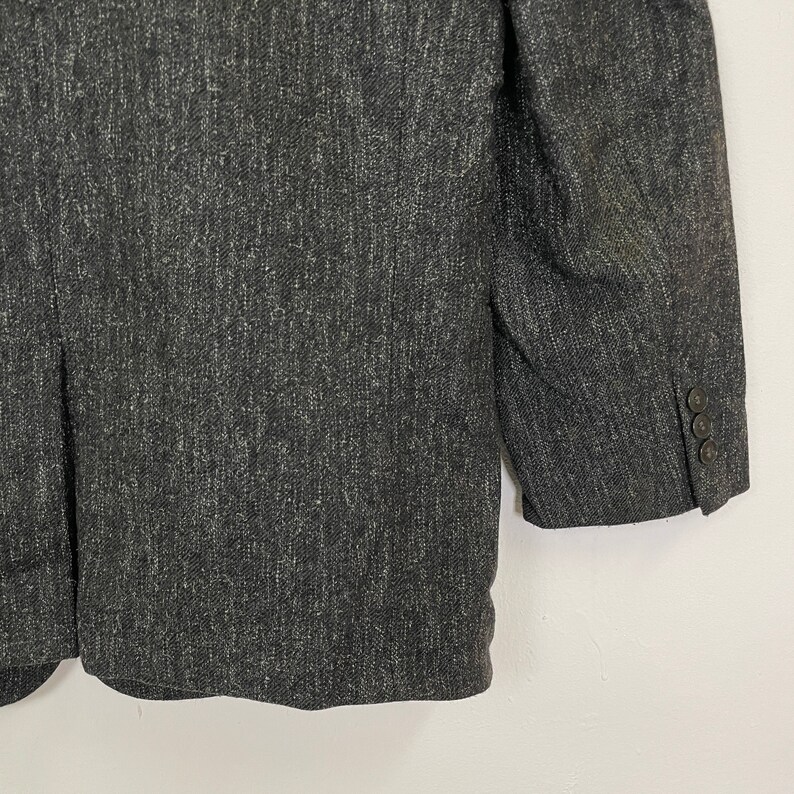 Op de afbeelding: Een donkergrijze blazer van een getextureerde stof. De blazer heeft een enkele knoopsluiting en twee zakken aan de voorkant.