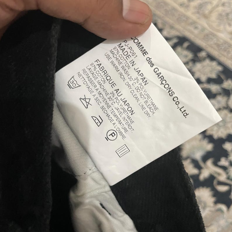 Puede incluir: Etiqueta de cuidado blanca con texto negro que detalla el contenido de la tela y las instrucciones de cuidado para una prenda. El texto dice "COMME des GAR&Ccedil;ONS Co., Ltd. PJ-P061 MADE IN JAPAN 97% COTTON 3% POLYURETHANE MACHINE WASH 30&deg;C. DO NOT BLEACH. USE WARM IRON. DRY CLEAN. LINE DRY. FABRIQUE AU JAPON 97% COTON 3% POLYURETHANE LAVAGE MACHINE 30&deg; C. JAVEL INTERDIT. REPASSER A MOYENNE TEMPERATURE. NETTOYAGE A SEC. SECHAGE A L'OMBRE."