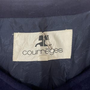May include: A white label with the text "Courr&egrave;ges" in black lettering. The label is attached to a dark blue fabric with a pin.