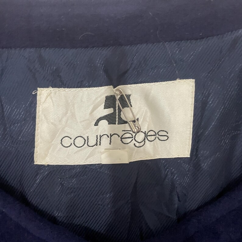 May include: A white label with the text "Courr&egrave;ges" in black lettering. The label is attached to a dark blue fabric with a pin.